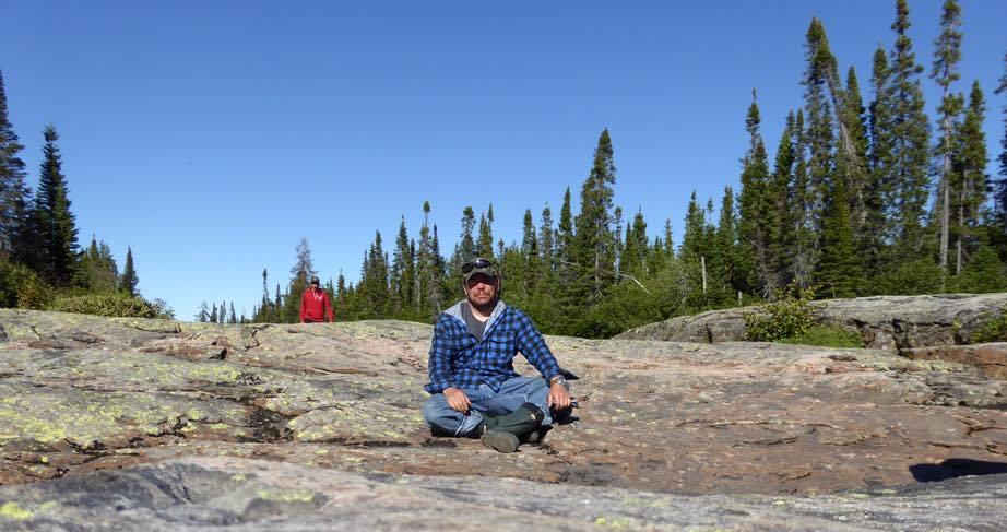 Homme en chemise à carreaux bleue et jeans assis sur un gros rocher.