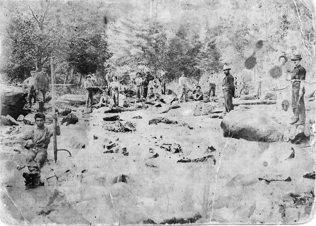 Photographie ancienne en noir et blanc présentant une douzaine de bûcherons en pleine forêt. À gauche, un homme est assis sur un rocher, et s’appuie sur un tourne-bille.