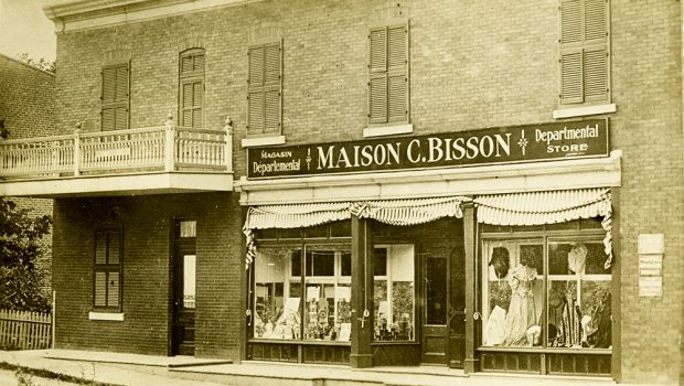Photographie d’époque en teinte sépia présentant la devanture d’un magasin départemental situé au rez-de-chaussée d’un bâtiment. Sur l’écriteau, on peut y lire : Maison C. Bisson.