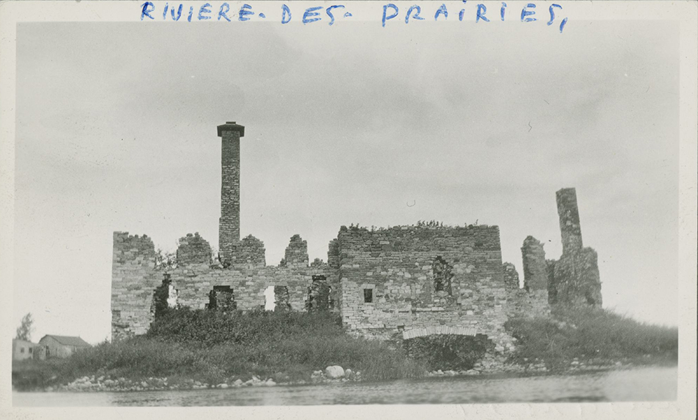 Photographie d’époque en noir et blanc présentant des ruines en pierre sur le bord d’une rive. Il s’agit d’un ancien moulin qui a été détruit. Sur la bordure blanche supérieure, il est écrit à la main : Rivière-des-Prairies.