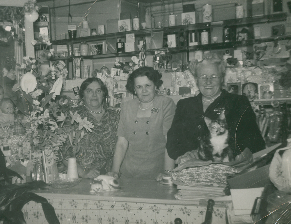 Photographie en noir et blanc présentant trois femmes derrière un kiosque à l’intérieur d’un magasin. 
