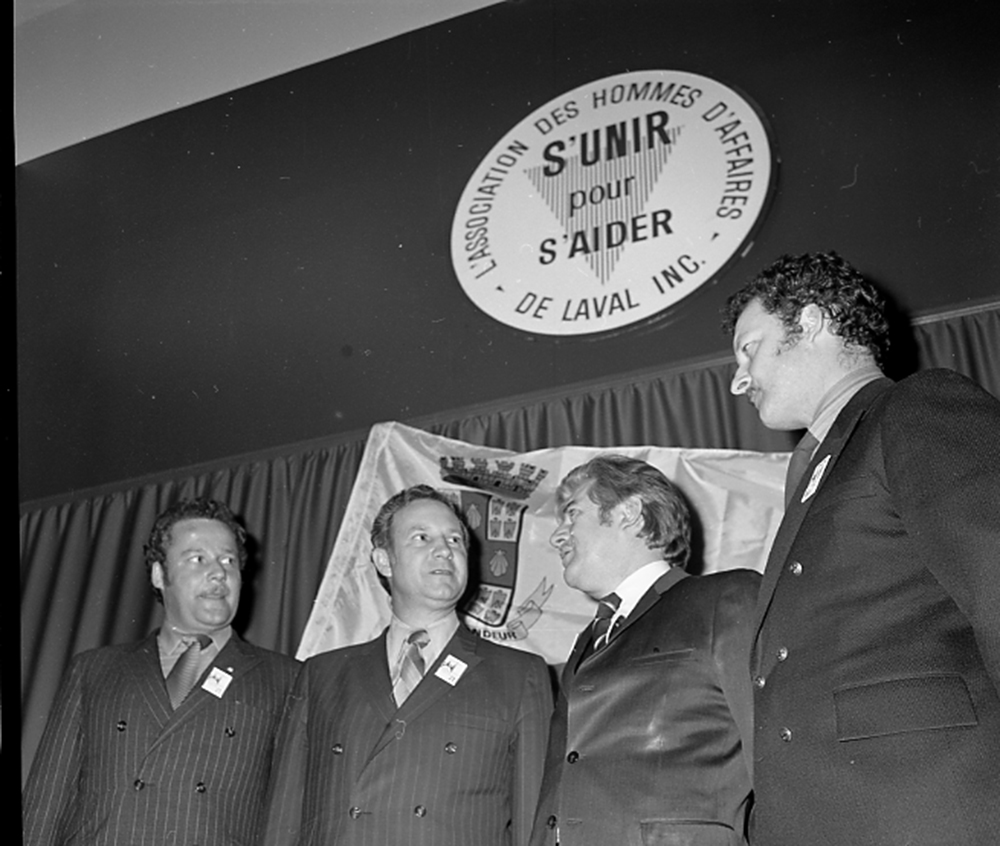 Photographie en noir et blanc de quatre hommes vêtus en complets noirs, qui semblent discuter entre eux. En haut sur le mur, on peut lire sur un écusson : « L’association des hommes d’affaires de Laval Inc. S’unir pour s’aider. »