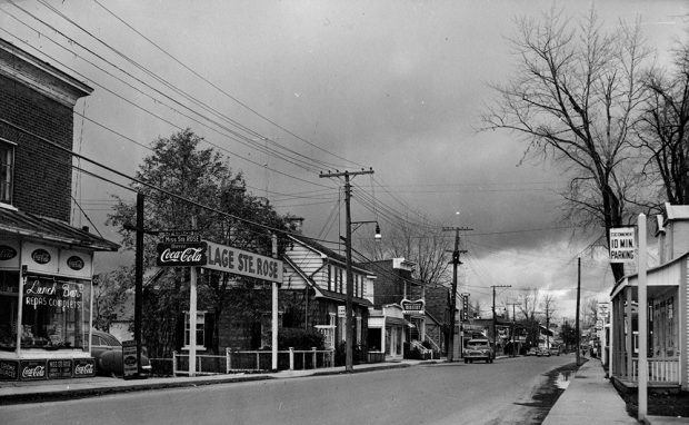 Photographie en noir et blanc montrant une rue commerciale dans le quartier de Sainte-Rose. On voit quelques magasins, des voitures stationnées sur le bord de la rue, et l’entrée vers la plage de Sainte-Rose.