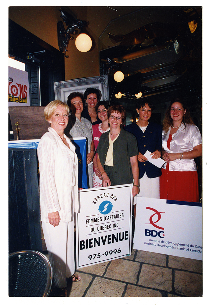 Photographie en couleur d’un regroupement de sept femmes dans une salle sombre. On peut lire sur deux pancartes mises en premier plan : « Réseau des femmes d’affaires du Québec Inc » et « Banque de développement du Canada ».