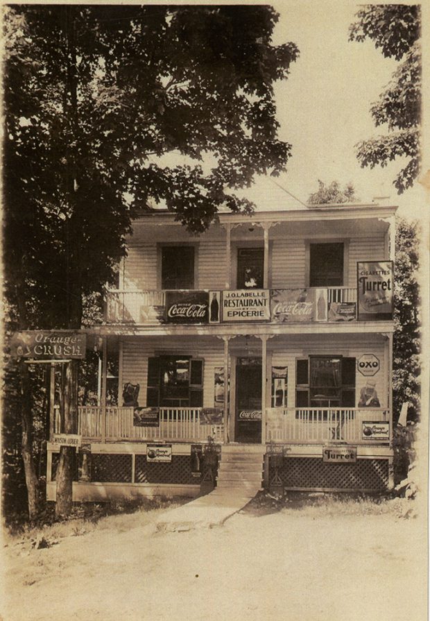 Photographie en teinte sépia d’une épicerie locale. Sur l’écriteau du duplex, il est écrit : J.O. Labelle Restaurant Épicerie. Sur la façade, on peut voir quelques panneaux mettant certains produits, dont les boissons Coca-Cola et Orange-Crush, et les cigarettes Turret.