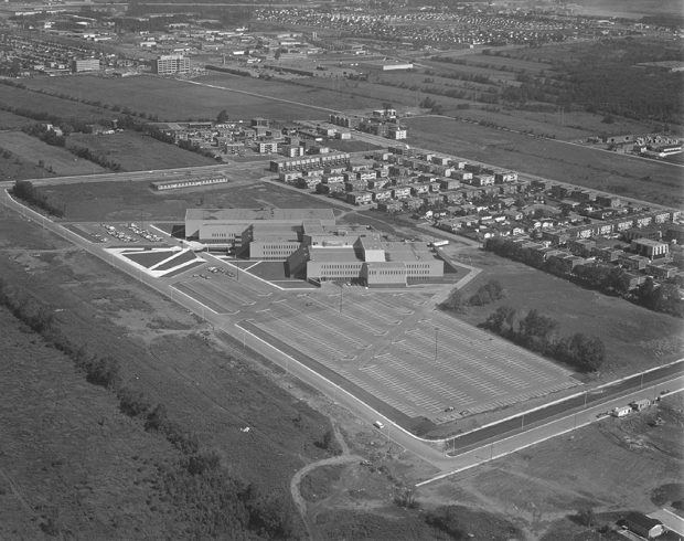 Photographie aérienne en noir et blanc du collège Montmorency. On y aperçoit en premier plan l’établissement avec un grand stationnement, entouré de champs et d’un petit quartier résidentiel. 