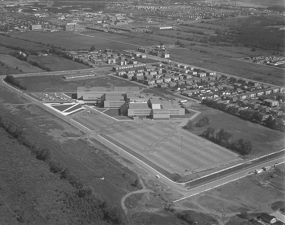 Photographie aérienne en noir et blanc du collège Montmorency. On y aperçoit en premier plan l’établissement avec un grand stationnement, entouré de champs et d’un petit quartier résidentiel. 
