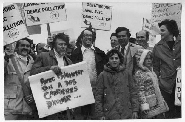 Photographie en noir et blanc montrant une dizaine de citoyens manifestant pour dénoncer la pollution par l’exploitation de la carrière Deluxe. En premier plan, on peut lire sur une pancarte : « Avec Paiement on a des carrières d’avenir… » 