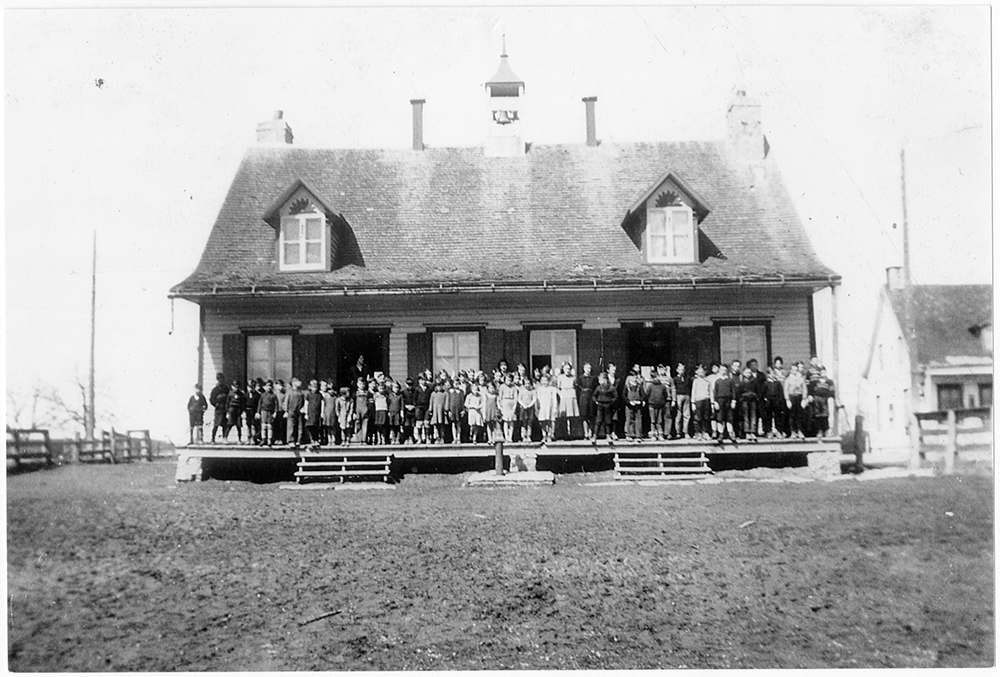Photographie en noir et blanc d’une petite école de rang. Une quarantaine d’élèves sont sortis sur le perron de la maison. 