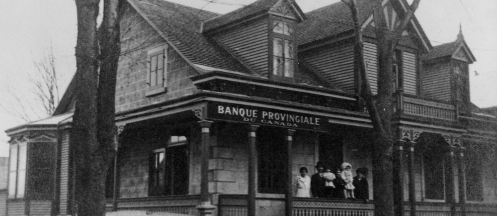 Photographie en noir et blanc d’une maison ancestrale. Il s’agit d’une succursale de la banque provinciale du Canada. Une famille est sur le perron. 