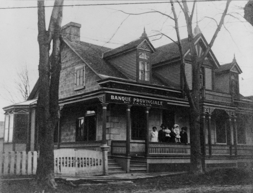 Photographie en noir et blanc d’une maison ancestrale. Il s’agit d’une succursale de la banque provinciale du Canada. Une famille est sur le perron. 