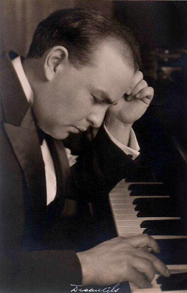 Photo noir et blanc d’Auguste Descarries au piano; il regarde pensivement le clavier avec une main au front et l’autre sur le clavier. 