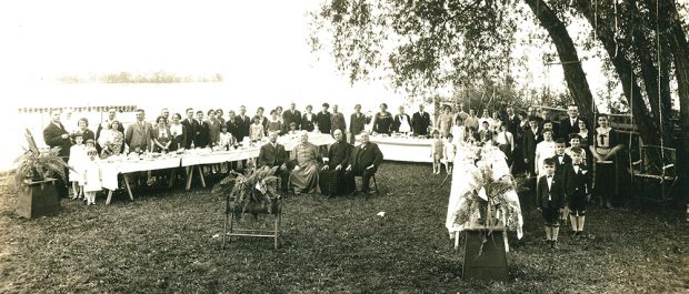 Photographie noir et blanc d'un grand groupe assemblé autour de table à l'extérieur. Il y a un étendue d'eau à l'arrière. Monseigneur Georges-Marie et son frère Armand sont devant le groupe avec 2 autres hommes.