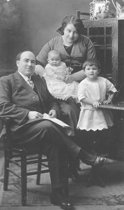 Black and white picture of Theophile LePailleur sitting at a work desk. His wife is behind him holding a child. Another child is standing next to them. They are in a studio setting.