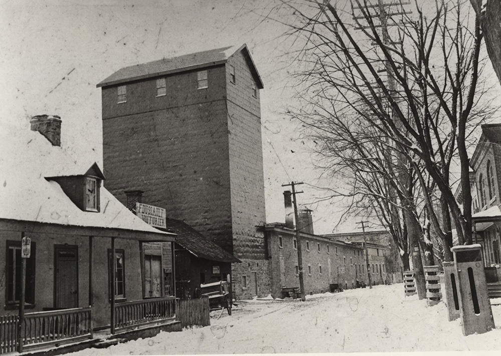 Photo noir et blanc d’un silo de plusieurs étages de haut et d’un long bâtiment de pierres avec une haute cheminée faisant partie des installations de la brasserie Dawes aux côtés de maisons résidentielles.