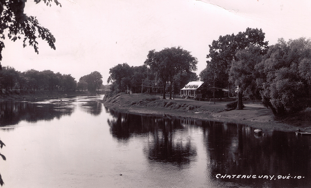 Photographie noir et blanc de la Maison Gravel. La rivière Châteauguay se trouve en avant-plan et plusieurs arbres en bordent les rives.