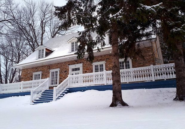Photo couleur de la Maison LePailleur prise l'hiver. Le balcon est bleu avec une balustrade blanche. Il y a deux gros conifères en premier plan.
