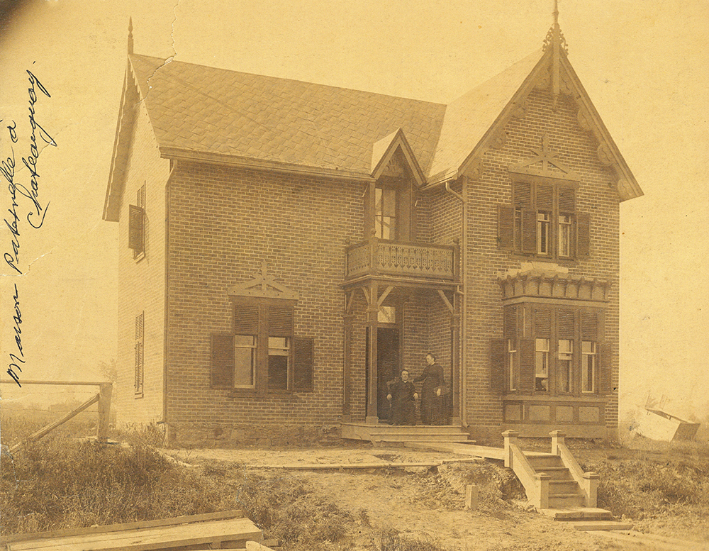 Photo sépia de Philomène Dalton sur le perron de la maison familiale avec son fil le curé Georges-Marie LePailleur. l y a une partie de clôture et un petit escalier en avant -plan mais le reste du paysage est plutôt vide.