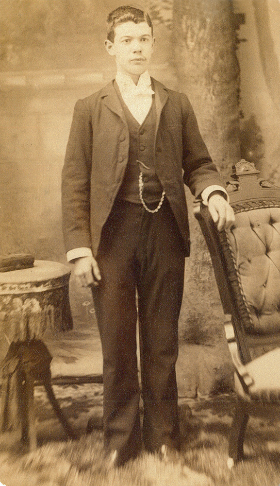 Photo sépia du jeune Armand LePailleur prenant la pose dans un studio photo. Il est debout sur un tapis de fourrure. Il porte un habit et une montre de poche.