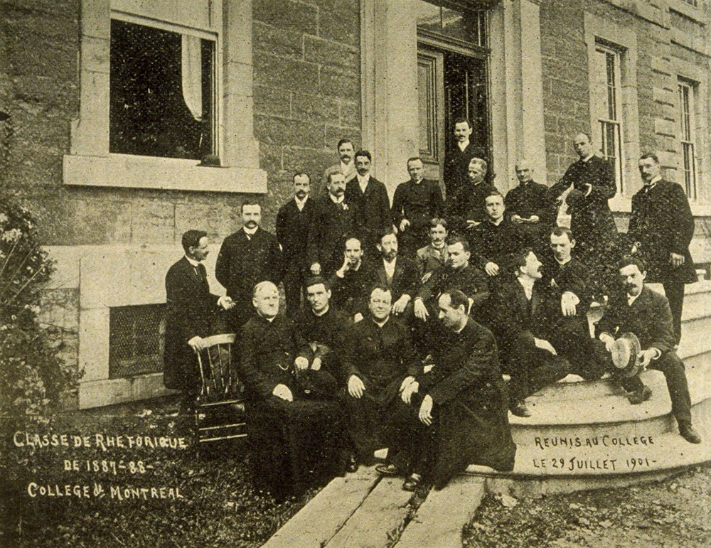 Photographie noir et blanc d'un groupe d'une vingtaine d'hommes devant le Collège de Montréal.  Quelques-uns portent la soutane, les autres sont en habit. Ceux dans les deux premières rangées sont assis, les autres sont debout.