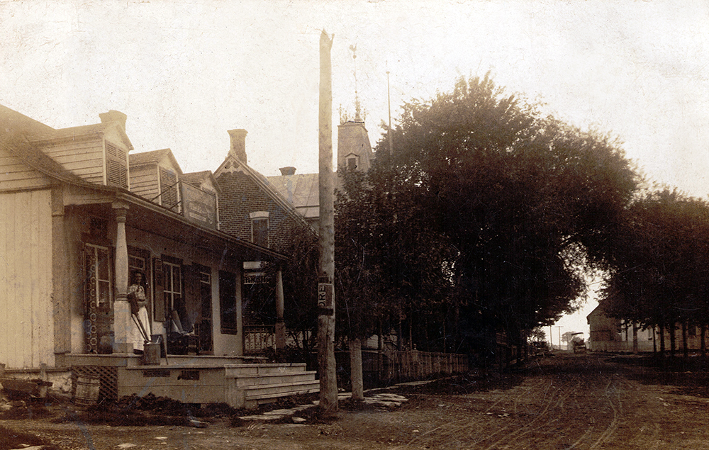 Photo noir et blanc de la rue Principale de Châteauguay. La route est en terre et des maison sont sur le côté droit. Une dame en robe longue est sur la galerie.