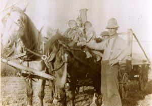 Three children on farm wagon horse beside 2 men.
