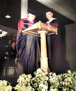 Deux hommes portant toge et mortier académique debout à un podium avec fleurs dans l'avant-plan