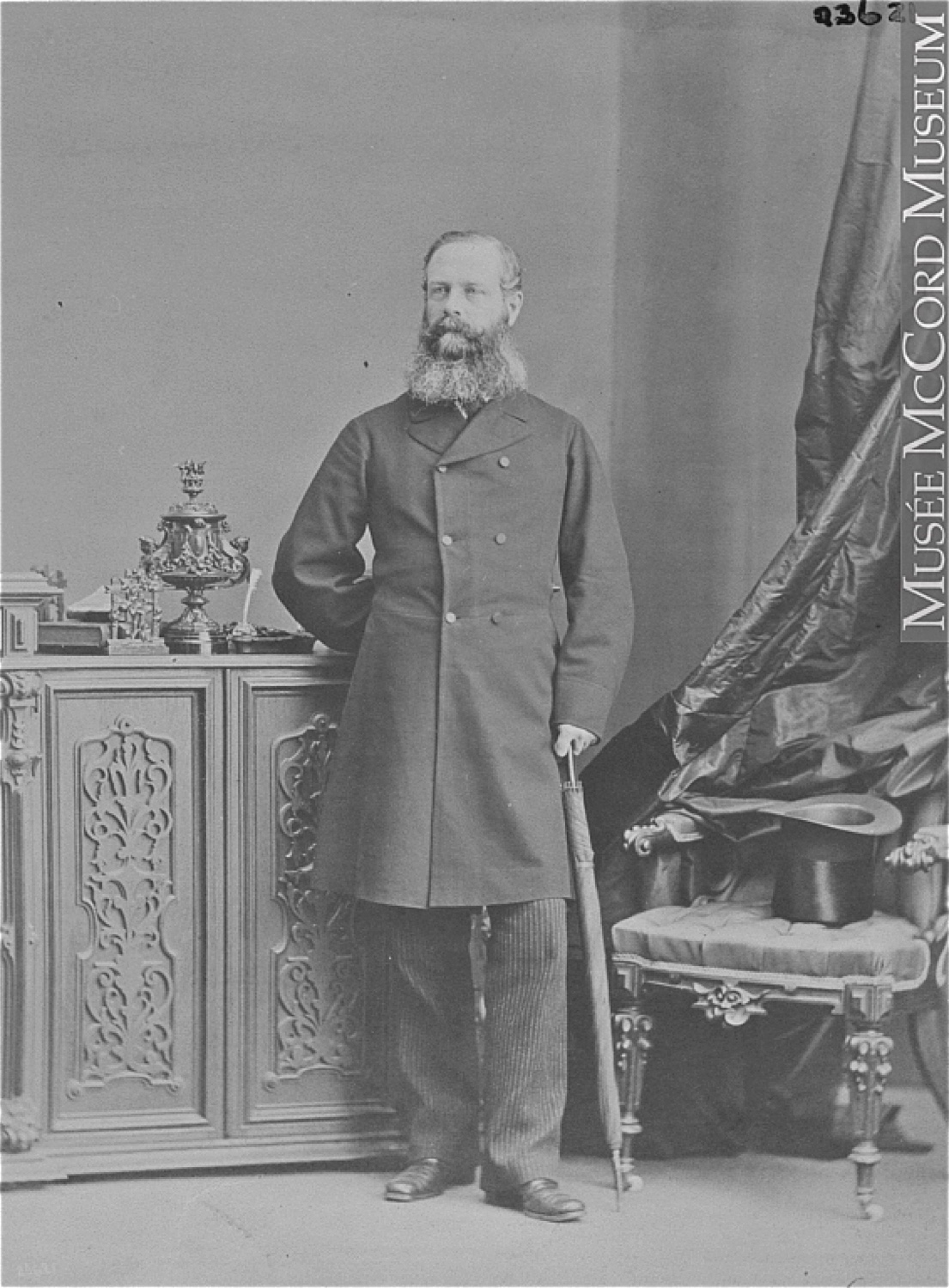 Photographie noir et blanc d'un homme (Lord Monck) debout, s’appuyant sur un parapluie replié, sa main droite derrière son dos, posant à côté d’une chaise sur laquelle il a déposé son haut-de-forme. Il affiche une barbe fournie et porte un pardessus de laine à double boutonnage.