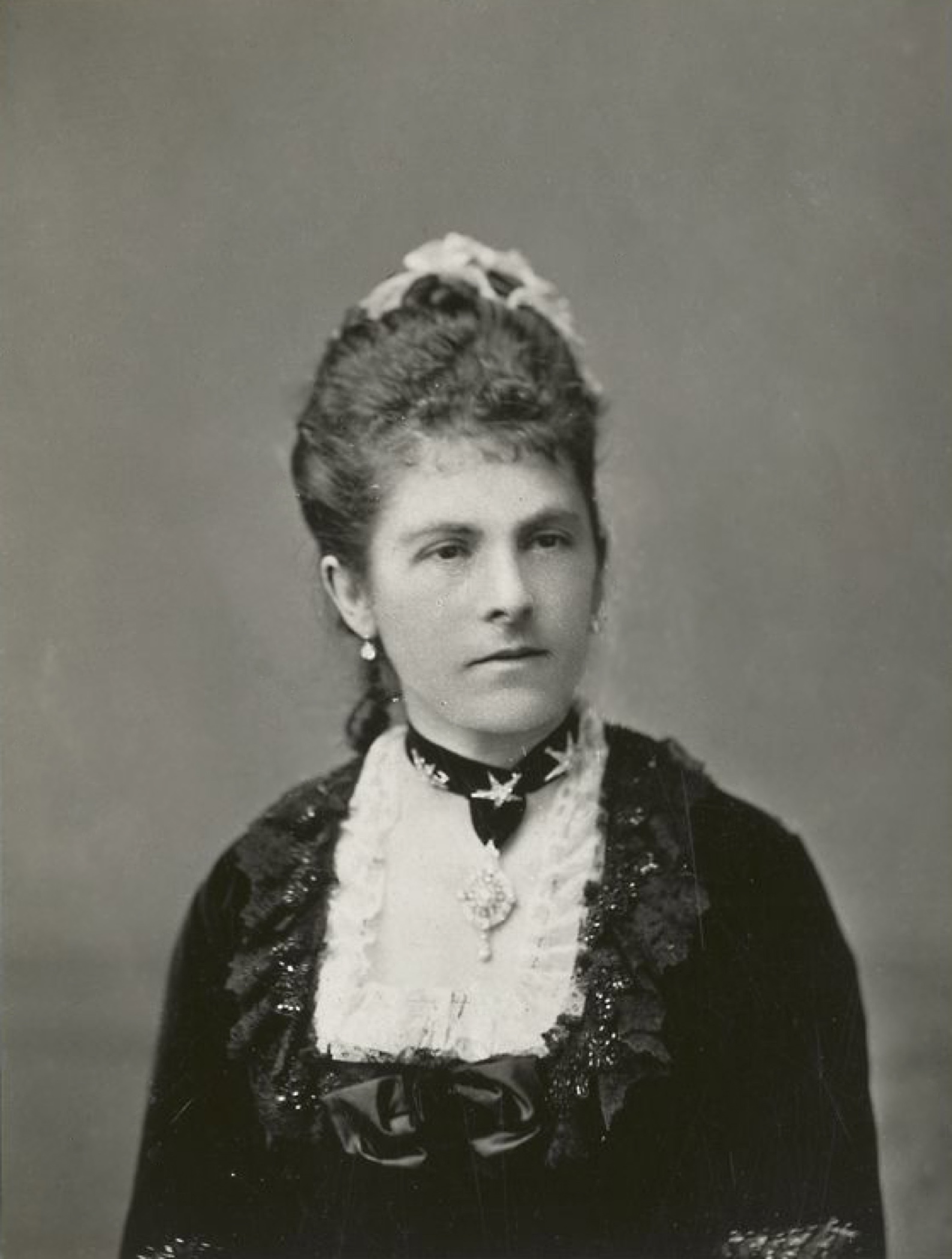 Photographie noir et blanc plan buste d'une femme (Lady Dufferin) dont les cheveux ont été coiffés; la dame est vêtue d'une robe de cérémonie à corsage en dentelle carré, parée d’un collier-ruban orné d’un bijou.
