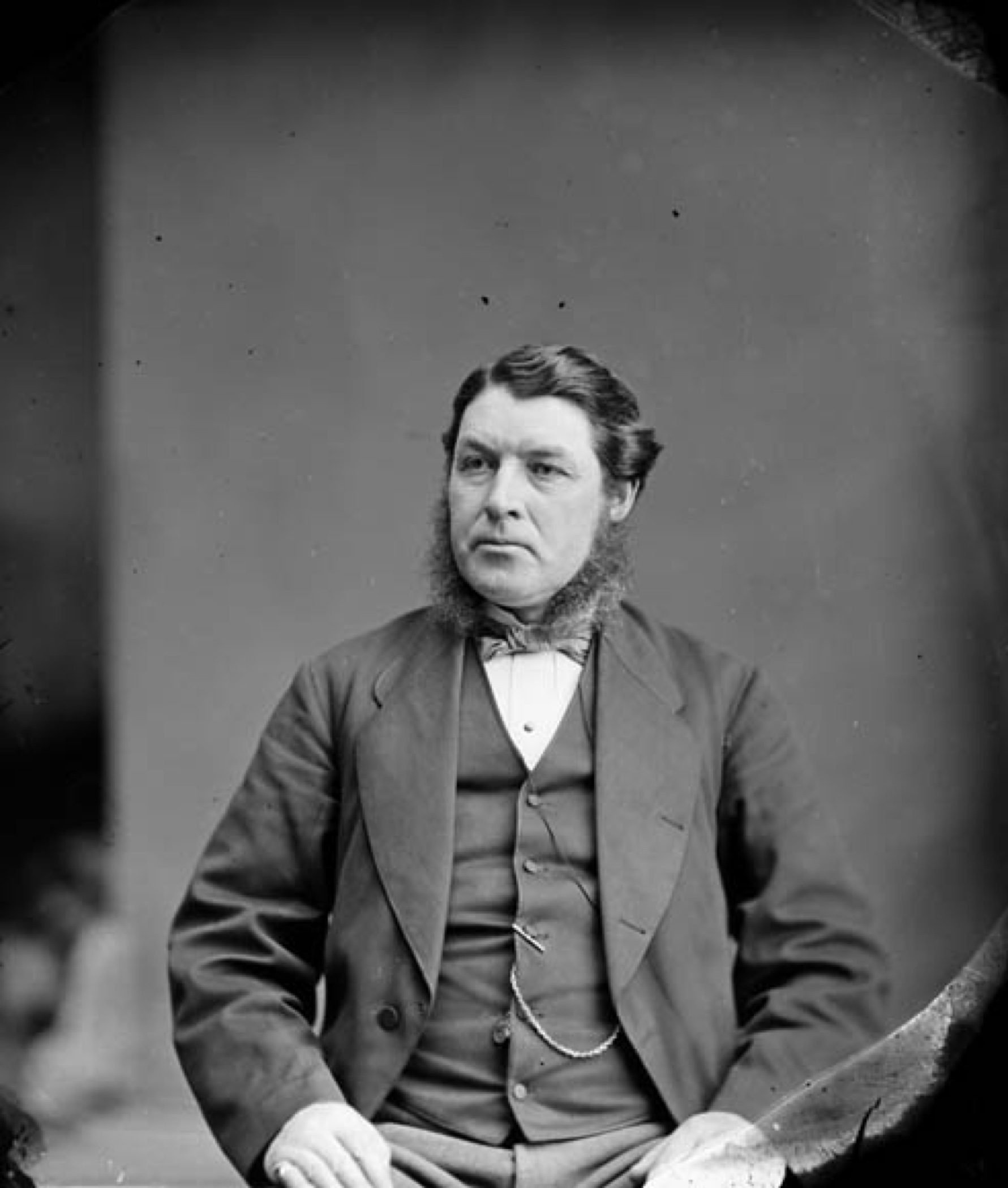 Photographie officielle en noir et blanc d'un homme (Sir Charles Tupper) à la chevelure noire et aux longs favoris, vêtu d’un costume avec veste, en position assise, les mains posées sur ses cuisses. On peut voir bien visiblement la chaine d’une montre sortant de son veston. Ses sourcils froncés et les coins de sa bouche abaissés lui donnent un air grave.