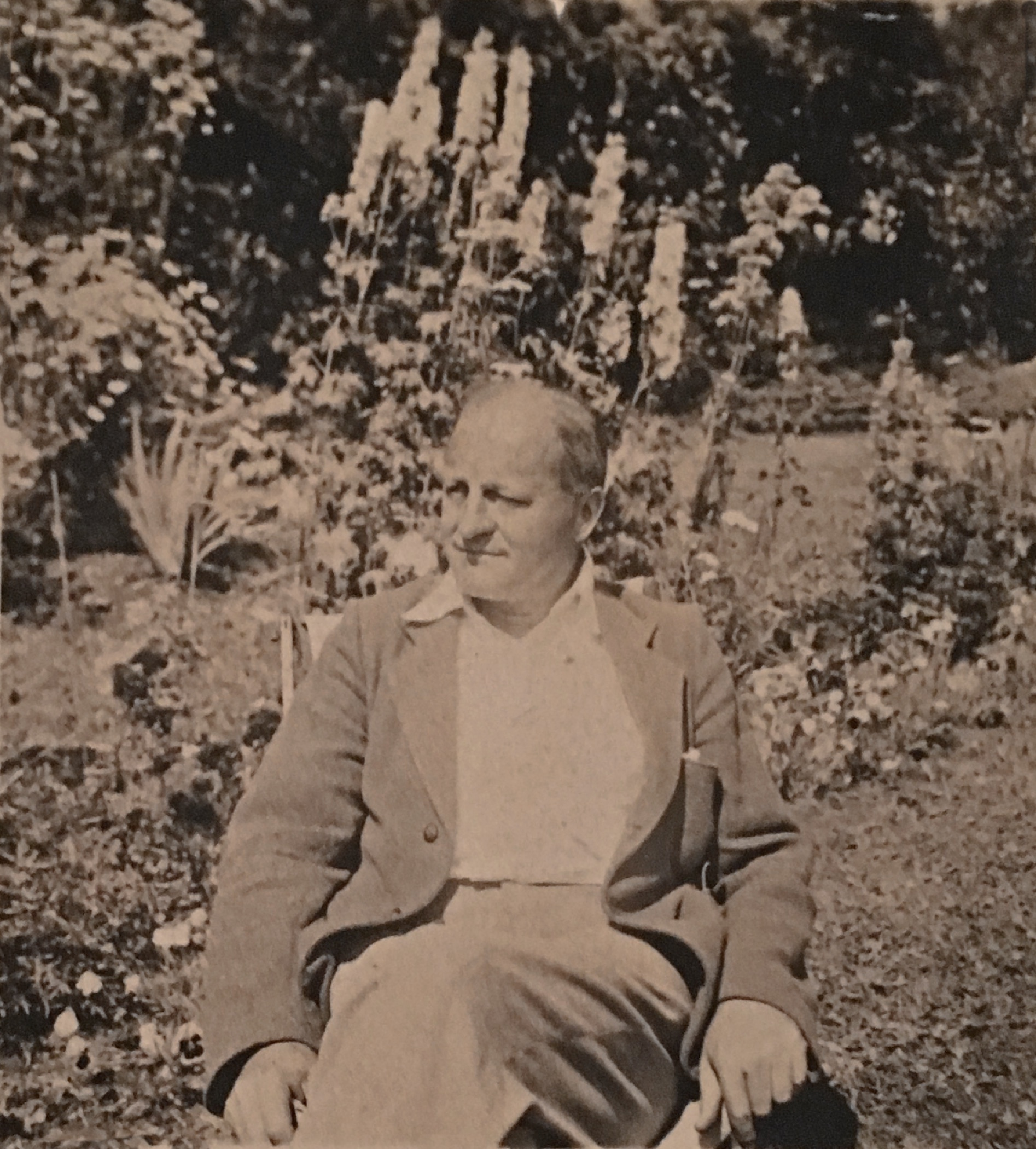 Photo aux tons sépia d'un homme d'âge moyen, Sir Herbert Symington, assis sur une chaise dans un jardin floral extérieur. Derrière lui se dressent de hauts delphiniums. 