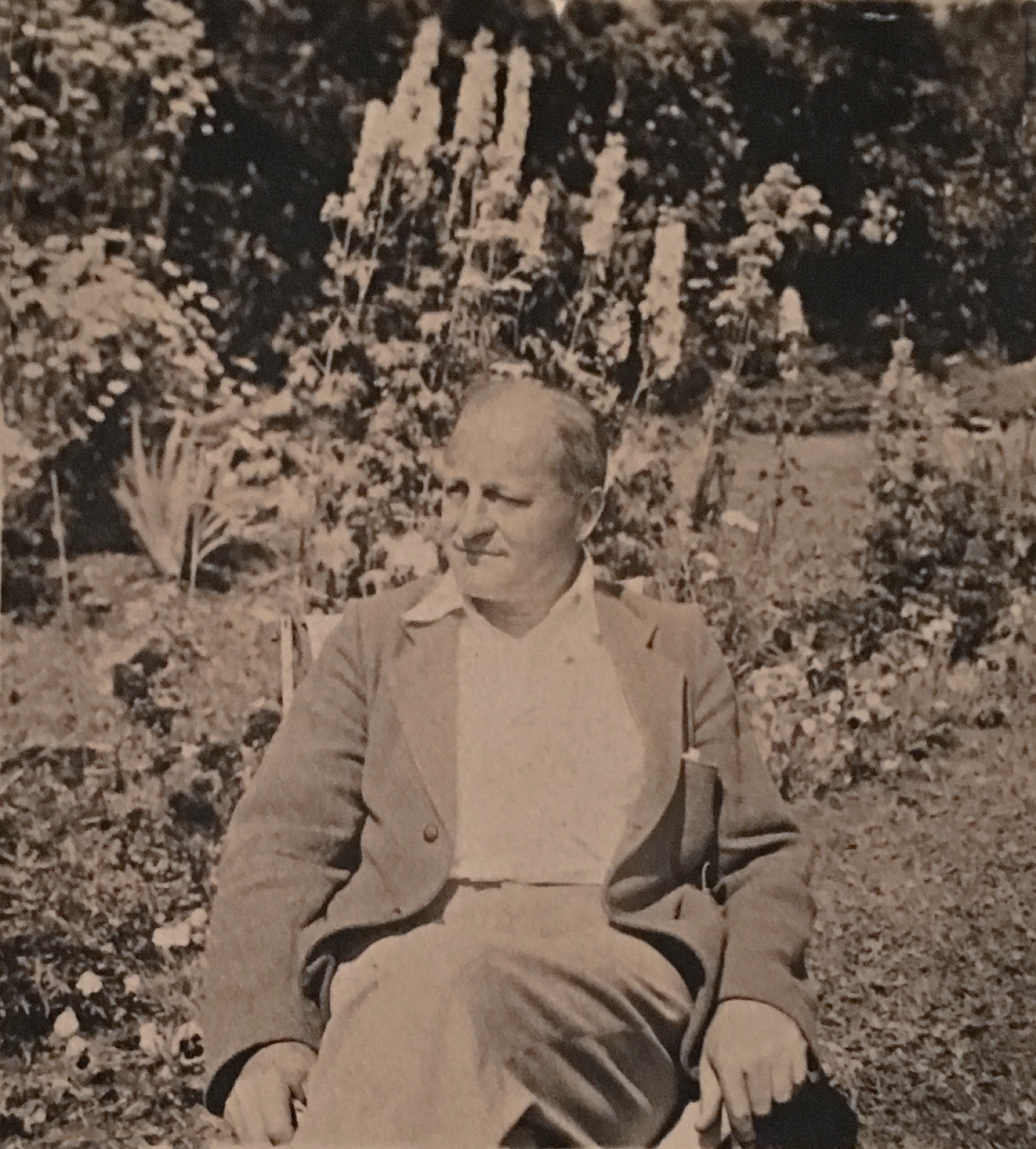 Photo aux tons sépia d'un homme d'âge moyen, Sir Herbert Symington, assis sur une chaise dans un jardin floral extérieur. Derrière lui se dressent de hauts delphiniums.
