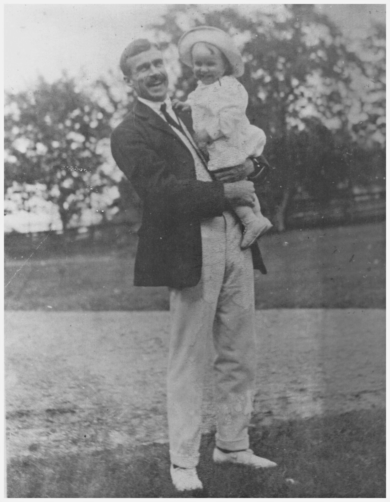 Photographie noir et blanc d'un homme de grande taille (Kenneth Molson), souriant, tenant dans ses bras un enfant en bas âge, lui aussi souriant, posant à l'extérieur sur une pelouse devant une rangée d’arbres en arrière-plan.