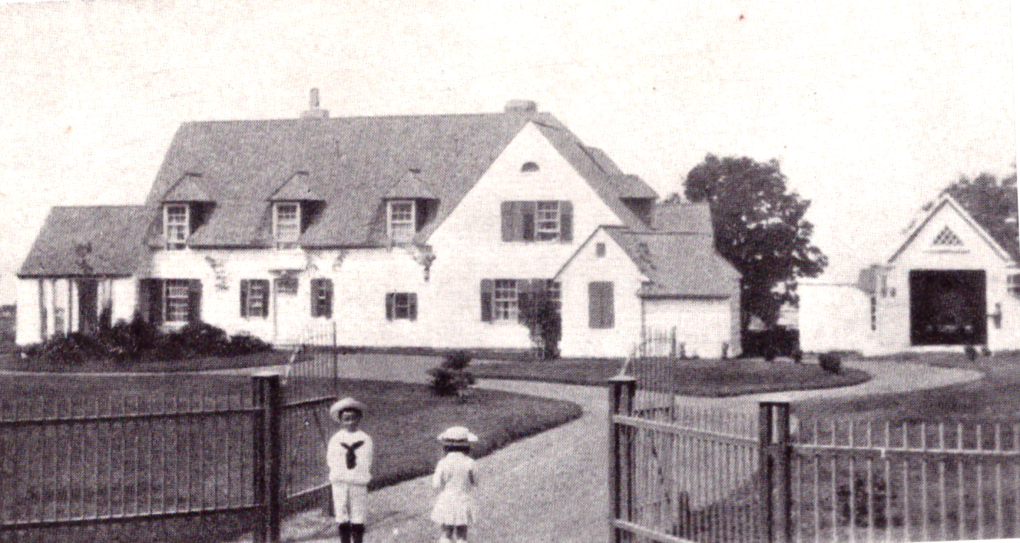 Photographie aux tons sépia d'une vaste maison (domaine de Kenneth Molson) entourée d'une clôture de fer avec portes; en avant-plan, on aperçoit deux jeunes enfants en costumes de matelots.