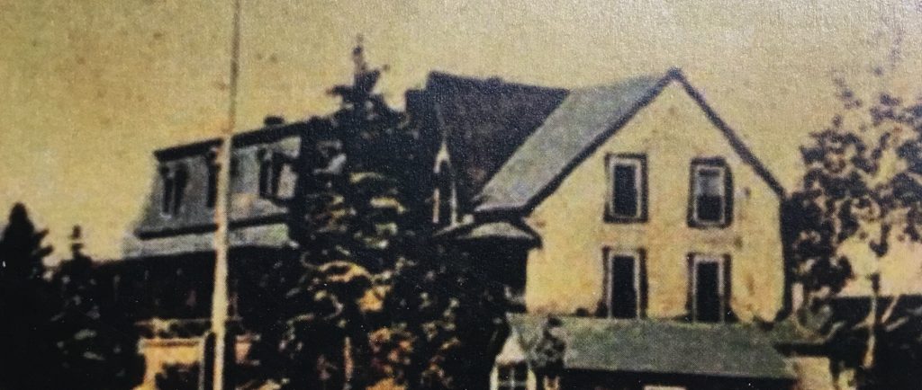 Photographie colorée à la main (à l'origine en noir et blanc) d'une grande maison (la villa Les Rochers) devant laquelle un mât de drapeau est érigé. Sur le côté de la propriété, on aperçoit une remise.