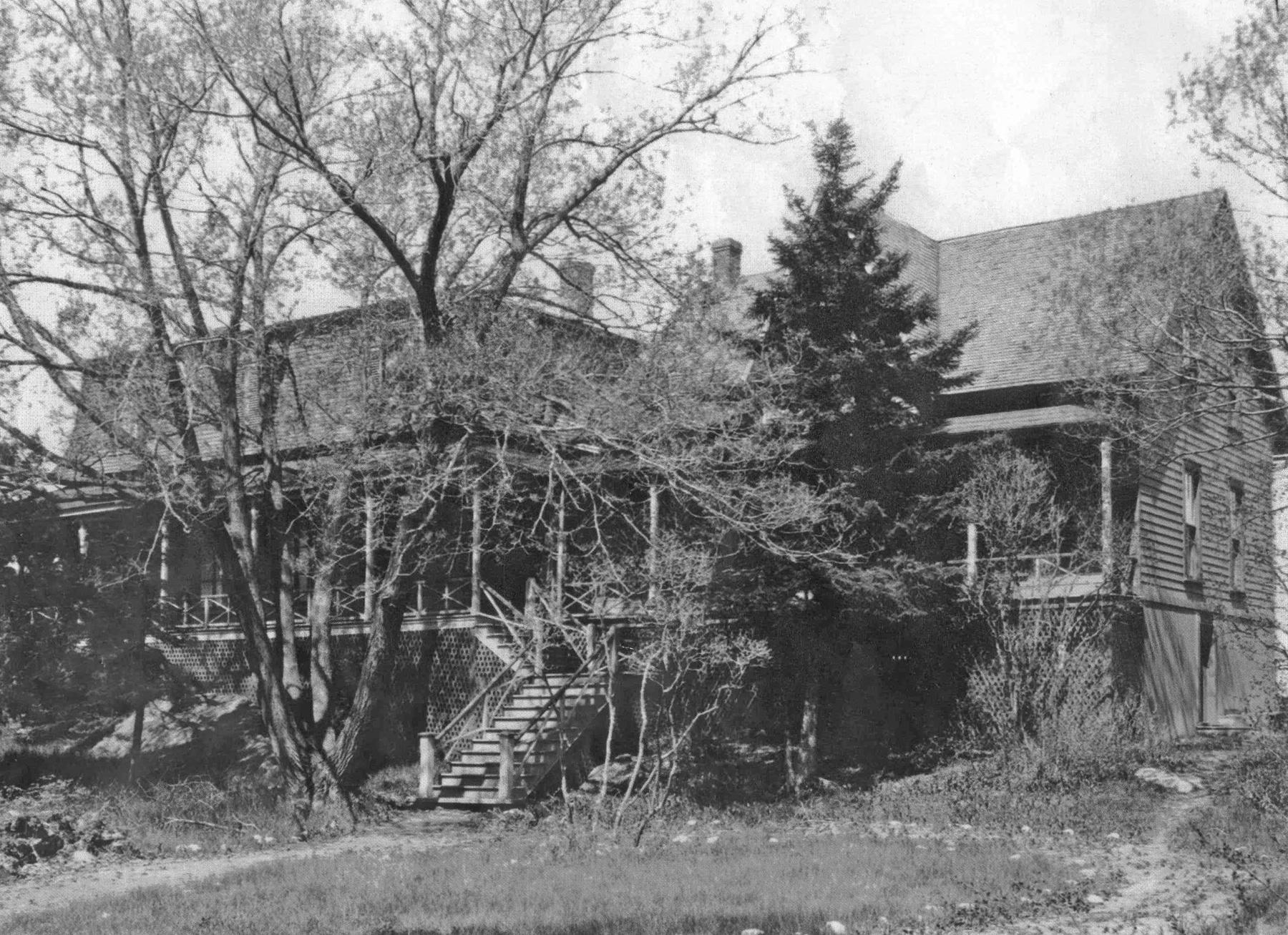 Photographie noir et blanc d'une grande maison (villa Les Rochers) dotée d'une large véranda avec escalier à l’avant, devant laquelle se dressent un arbre à feuillage caduque ainsi qu’un imposant conifère. 