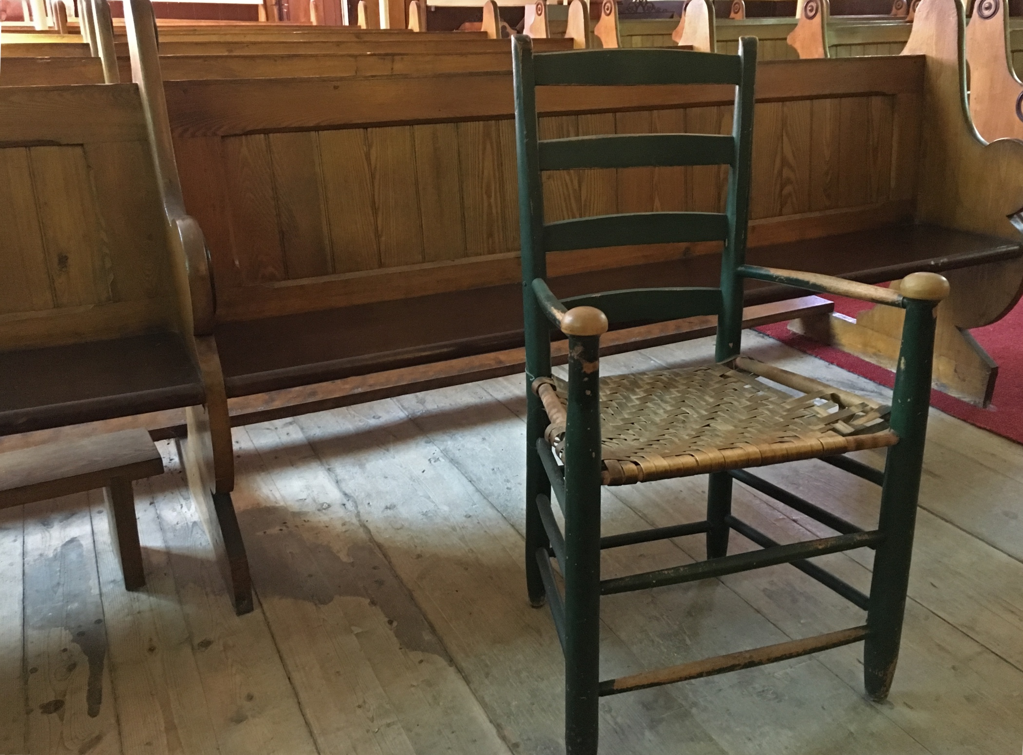 Photographie couleur d'une chaise en bois avec un siège à lattes, peinte en vert, dont on peut constater la vétusté par les lattes détachées du cadre sur un côté du siège ainsi que la peinture écaillée sur les appuie-mains des accoudoirs. La chaise se trouve devant une rangée de bancs d’église en bois. On aperçoit une surface du vieux plancher de bois franc brut.