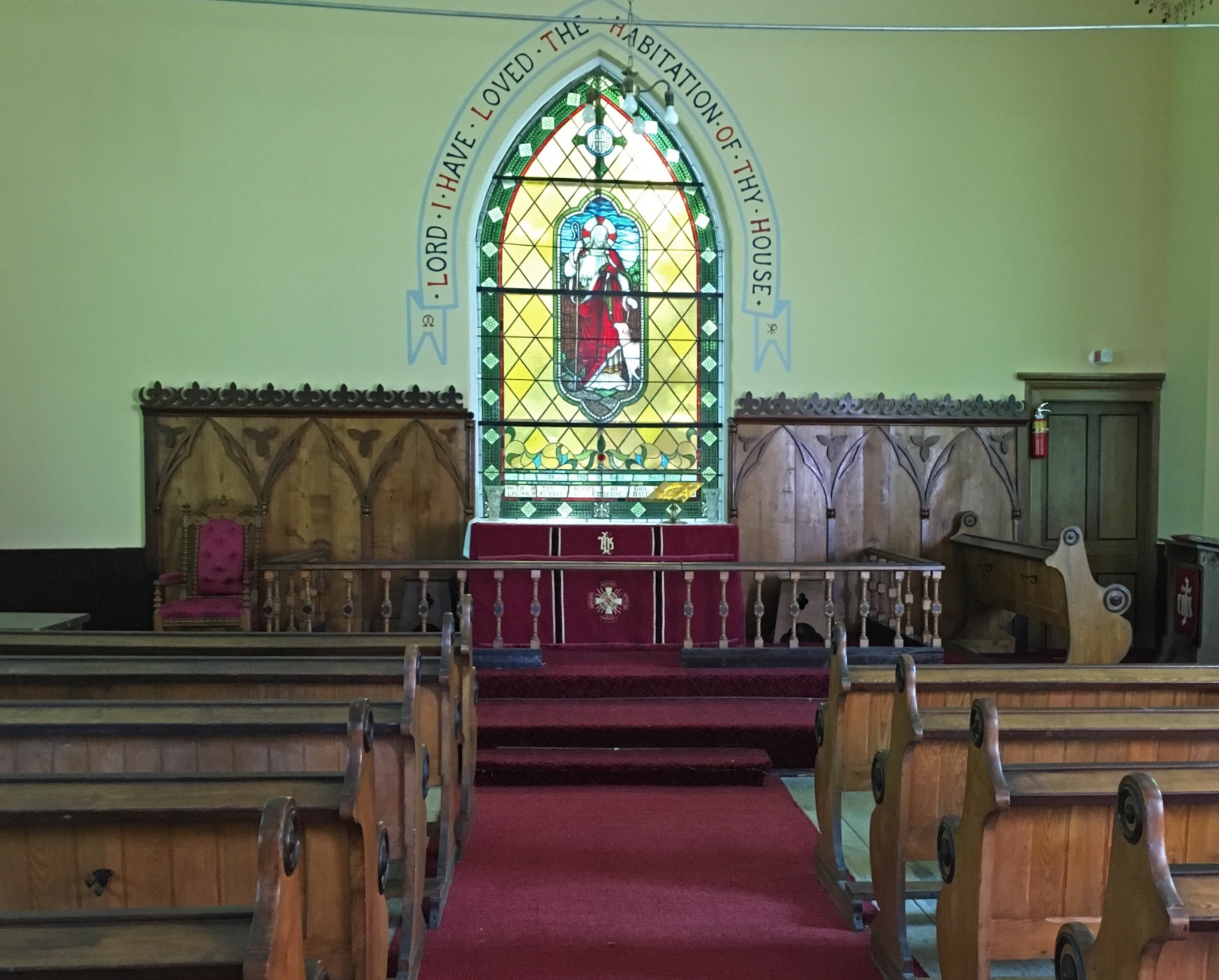 Photo couleur de l’intérieur de l'église anglicane de St. Bartholomew sur laquelle on aperçoit des rangées de bancs en bois faisant face à l’autel, lequel est orné de panneaux en bois fixés au mur sans apparat au centre duquel se trouve un vitrail.