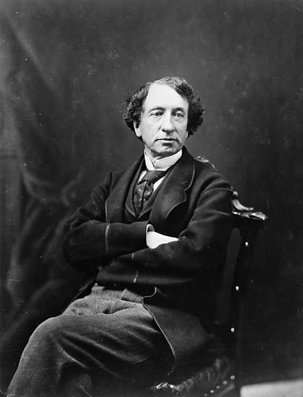 Portrait noir et blanc d'un homme d'âge moyen (Sir John A. Macdonald) à la chevelure noire, posant assis sur une chaise dans un studio, les bras croisés sur sa poitrine.