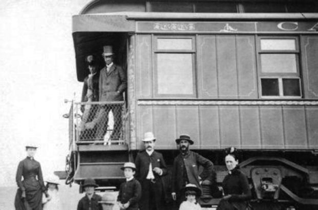 Photographie noir et blanc d'un couple (Sir John A. et Lady Macdonald) se tenant sur la plateforme d'un wagon de train, devant lequel pose un groupe de huit personnes, hommes, femmes et enfants. 