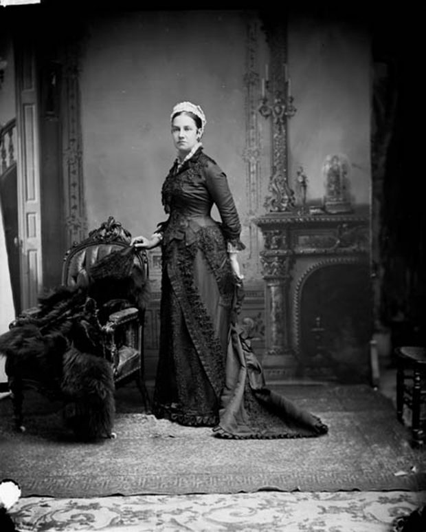 Photo noir et blanc d’une femme (Lady Agnes Macdonald) vêtue d'une longue robe noire, se tenant debout devant un foyer, à côté d'une chaise sur laquelle est déposé un manteau de fourrure. 