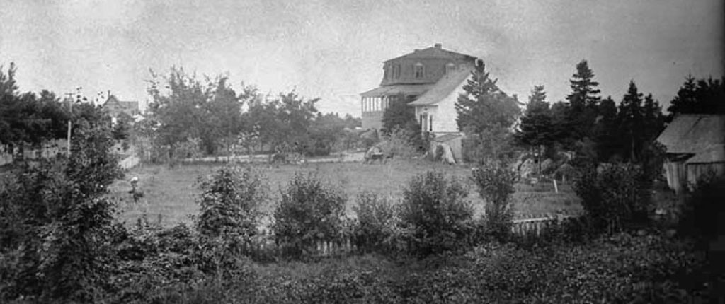 Photo noir et blanc de la villa Les Rochers v. 1883 et de son terrain bordé de conifères. Au premier plan à droite, on aperçoit un hangar en bois et en arrière-plan, la toiture en pointe d’un cottage voisin. 