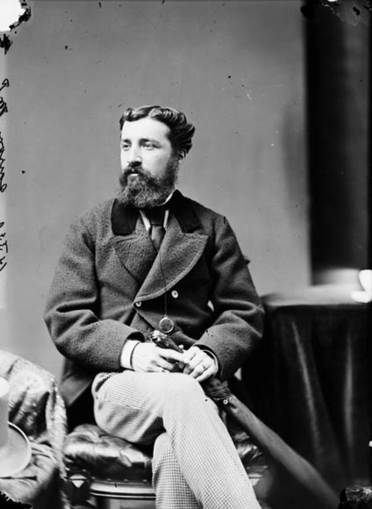 Photographie noir et blanc d'un jeune homme barbu (Sir Adolphe Caron) assis, tenant un parapluie fermé près de ses jambes croisées, son regard tourné vers la gauche. Il est vêtu d’un manteau de laine à double boutonnage et d’une cravate sombre. 