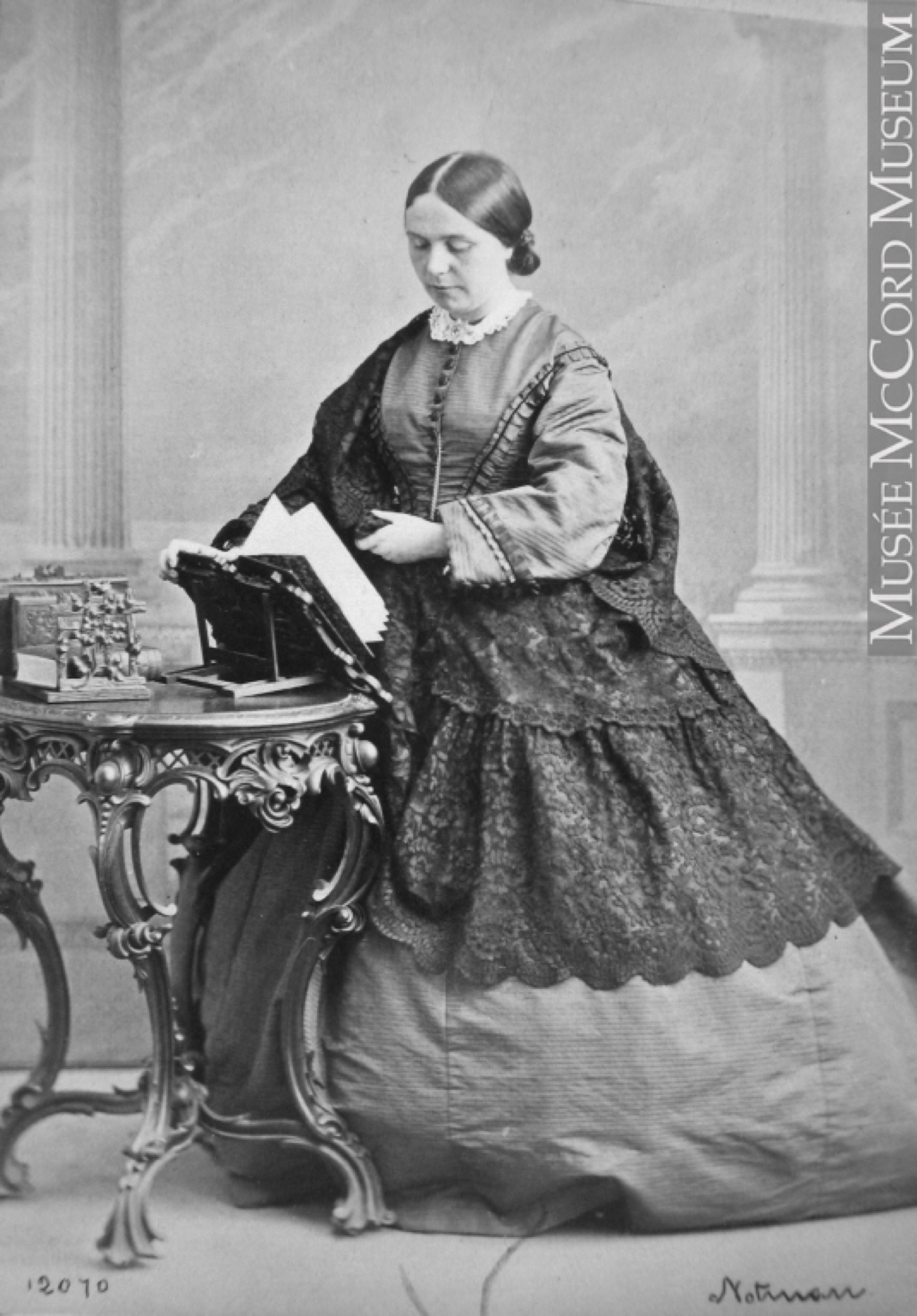 Photo noir et blanc d'une femme (Lady Frances Monck) se tenant debout, vêtue d’une large jupe longue drapée de dentelle, tournant les pages d'un livre déposé sur un lutrin devant elle. 
