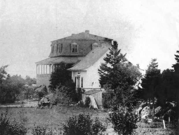 Photo noir et blanc d'une bâtisse (la villa Les Rochers) constituée de deux parties: une maison de ferme simple à charpente triangulaire, agrandie d’une annexe dotée d’un toit en mansarde. Des arbres et des arbustes entourent la propriété. 