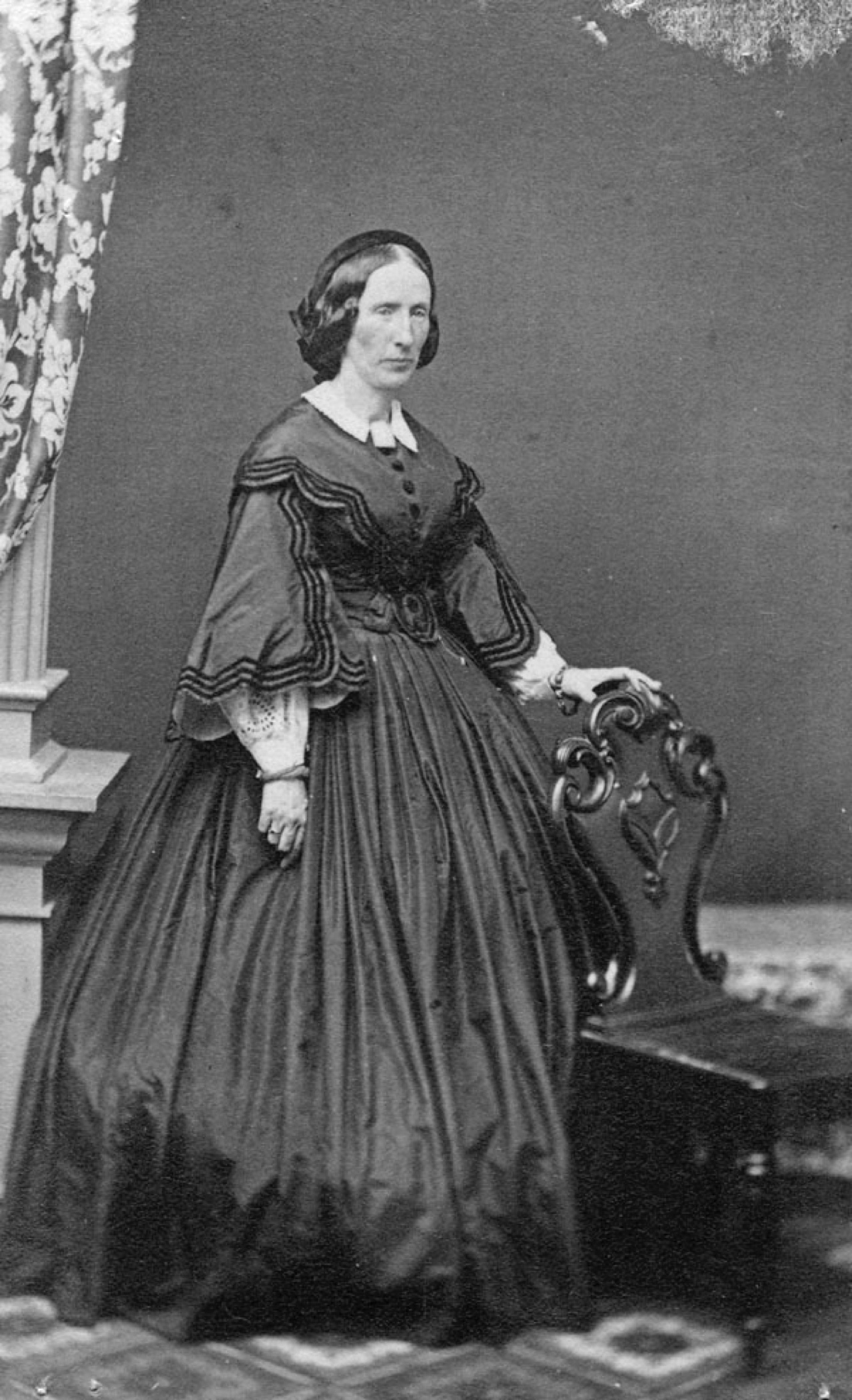 Portrait officiel en noir et blanc d'une femme (Louisa Macdonald) debout, vêtue d’une robe noire, sa main gauche posée sur le dossier d'une chaise. Ses cheveux noirs sont séparés par une raie au milieu et disposés à l'arrière. Son expression est lointaine et austère. 