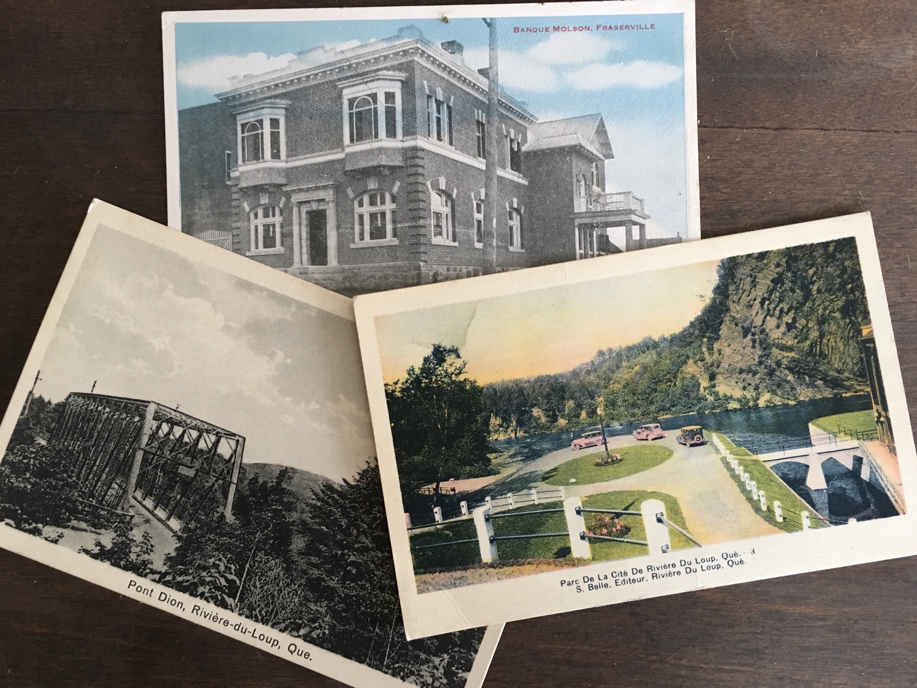 Photographie de trois cartes postales anciennes, coloriées à la main, se chevauchant sur une table, chacune illustrant différents paysages de Rivière-du-Loup. 