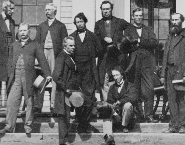 Photo noir et blanc de Sir John A. Macdonald assis sur les marches extérieures devant l’entrée de l’édifice Province House (Charlottetown), entouré des Pères de la Confédération canadienne. 