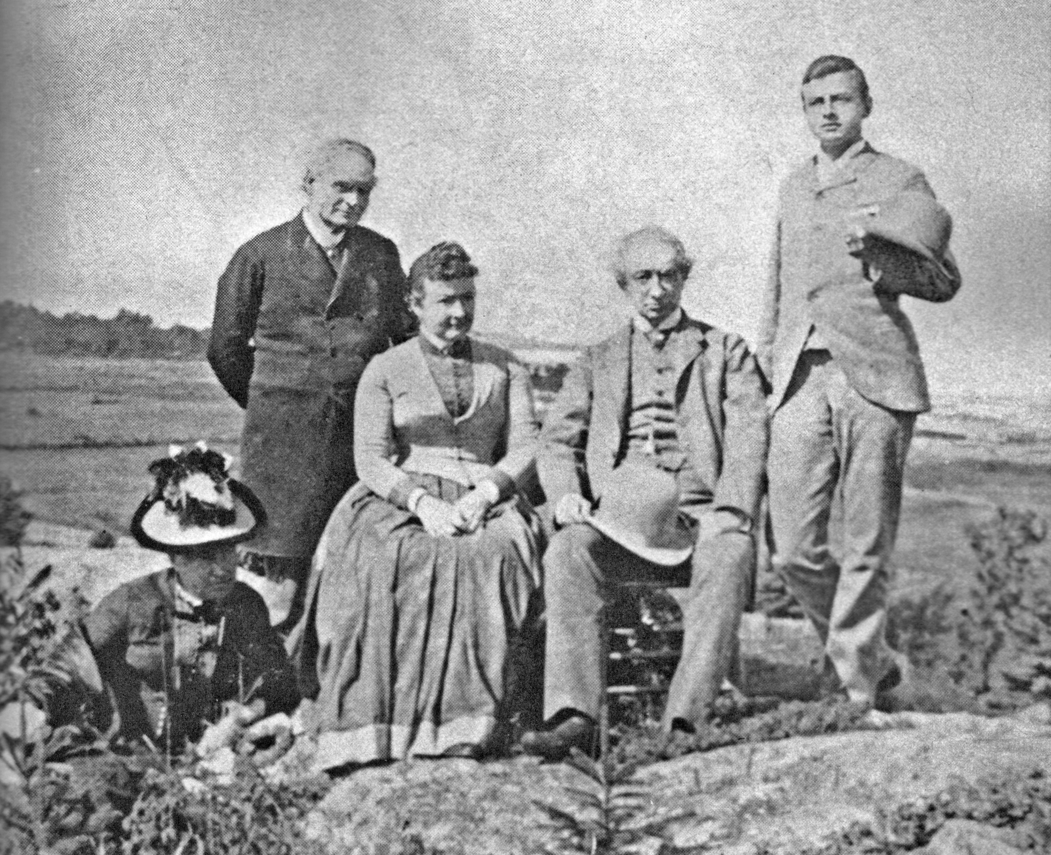 Photo granuleuse noir et blanc montrant cinq adultes (le couple Macdonald et les Tilleys) en positions debout et assise sur un sol rocheux, posant devant le fleuve St-Laurent en arrière-plan.