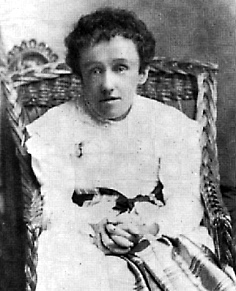 Photographie noir et blanc d'une jeune femme mince (Mary Macdonald) légèrement penchée vers l’avant, assise dans un fauteuil en osier, les mains jointes sur ses genoux. Elle porte une robe blanche avec une ceinture noire; ses cheveux sont sombres et son regard est vide. 